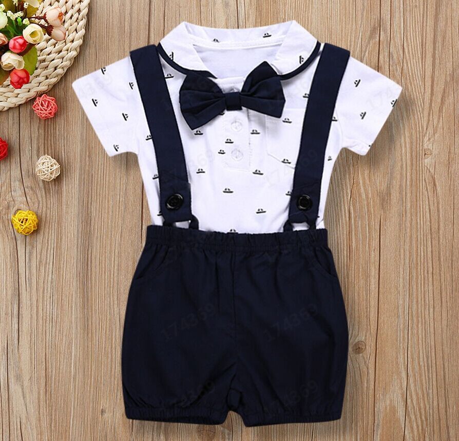Children's outfit with white shirt, black bow tie, suspenders, and dark shorts on a wooden surface.