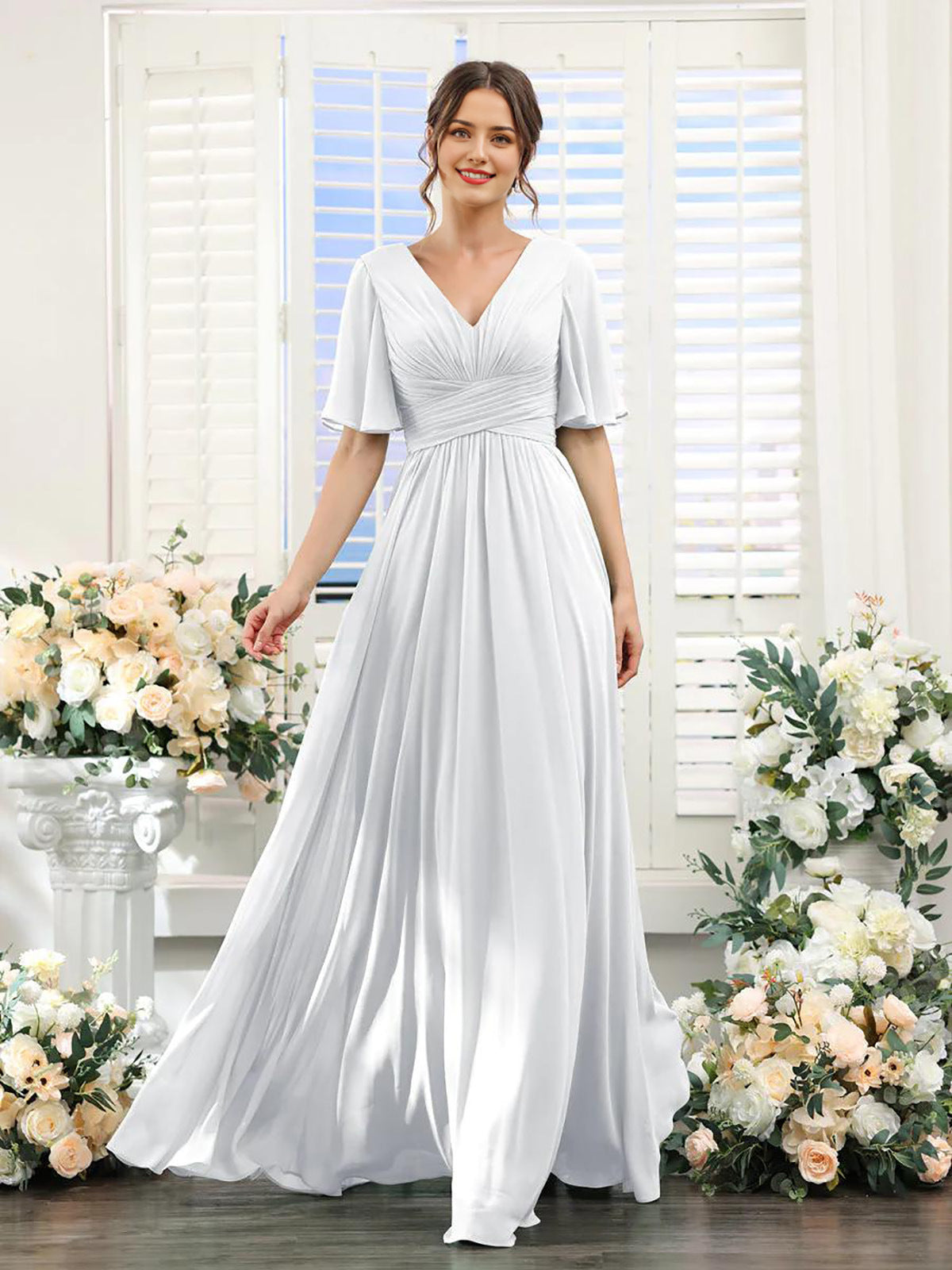 Woman wearing a white dress standing in front of floral decorations and a window with blinds.