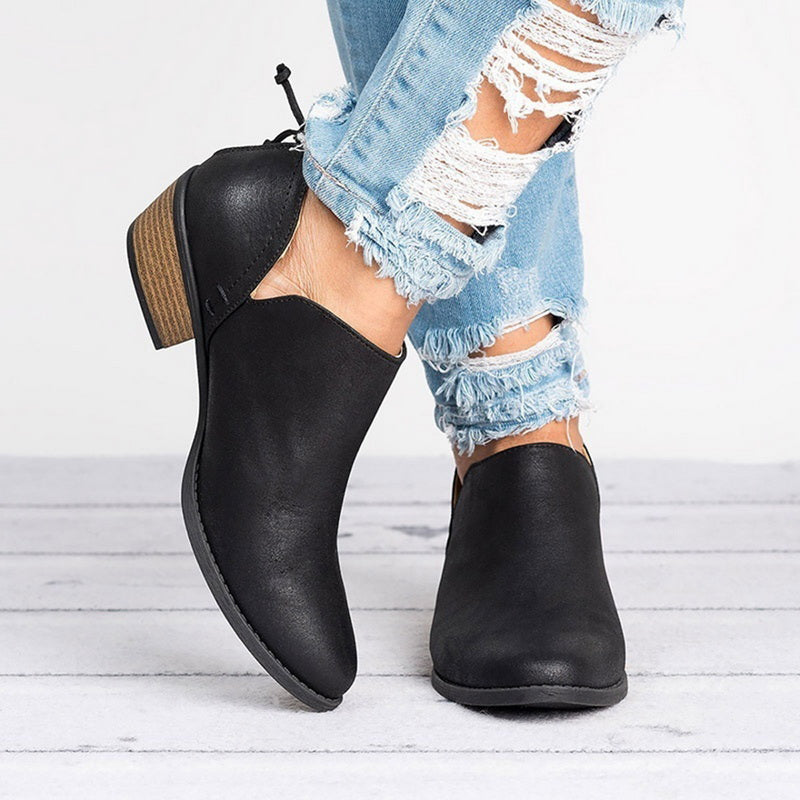 Black ankle boots worn with ripped denim jeans on a light wooden floor.
