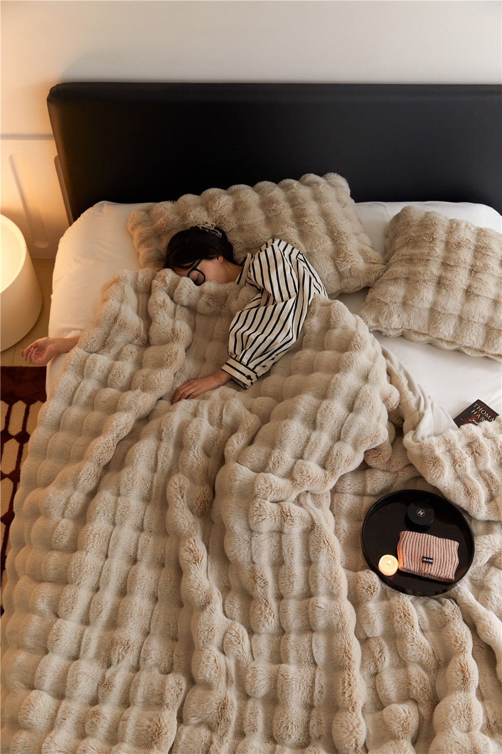 Person lying on a bed with a fluffy beige blanket, surrounded by soft lighting.