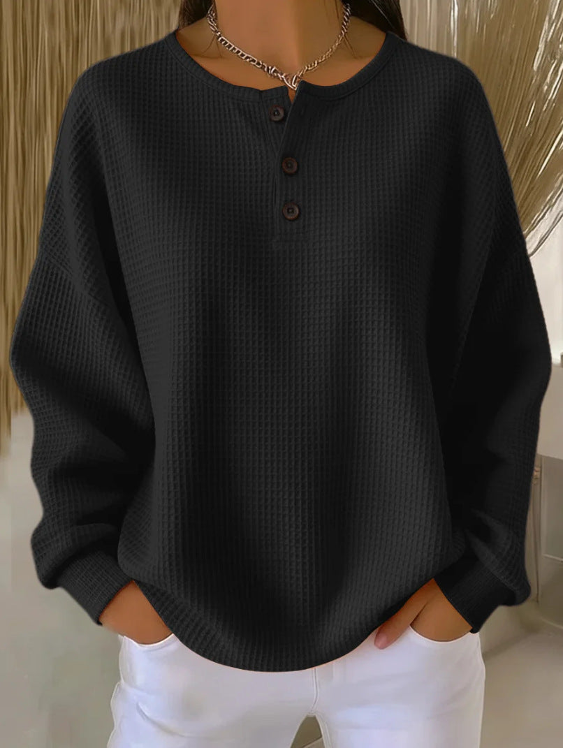 Person wearing a black textured sweater with buttons and white pants against a neutral background