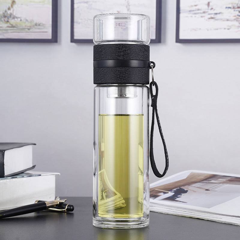 Clear glass water bottle with black lid and handle on a desk with books and photos in the background