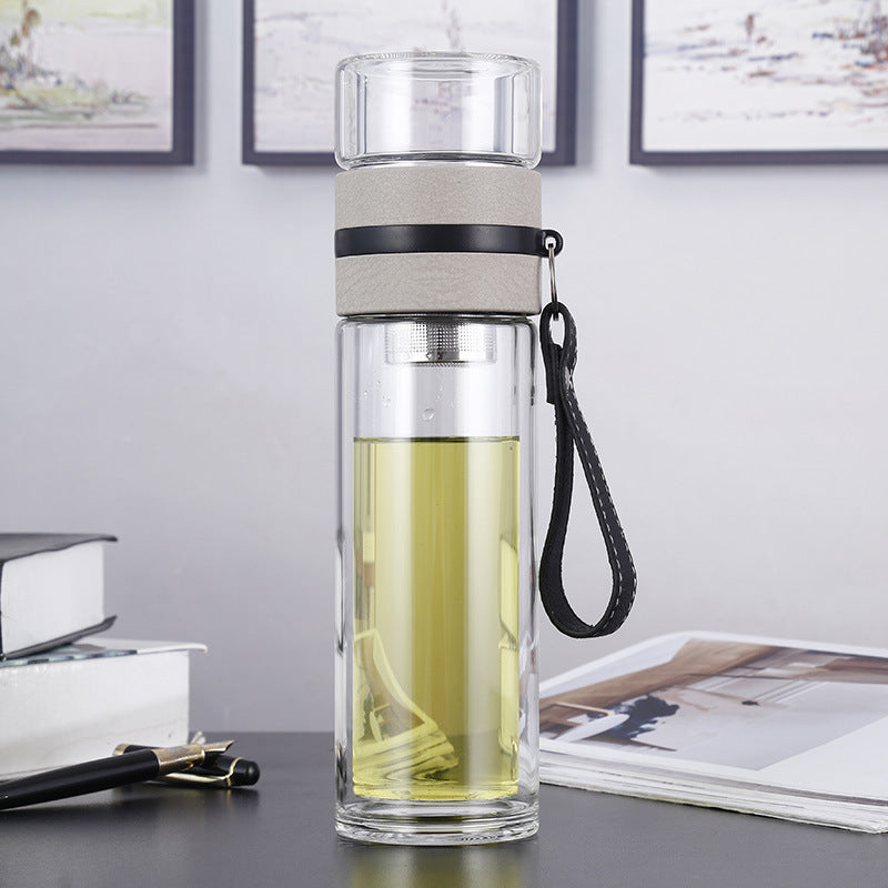 Clear glass water bottle with a gray cap and black strap on a desk with books and a pen.