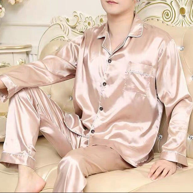 Man wearing pink satin pajamas sitting on a decorative couch with floral arrangements in the background.