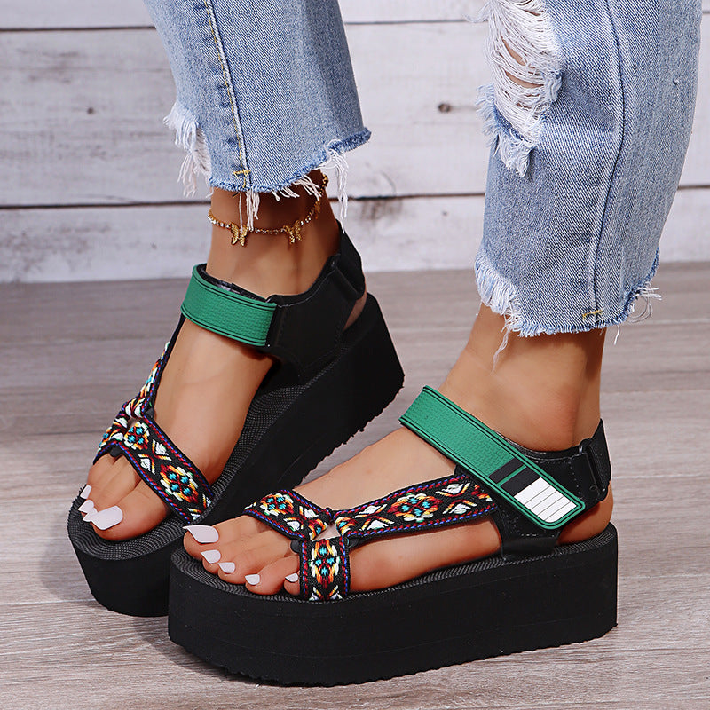 Black platform sandals with colorful straps worn by a person on a wooden floor.