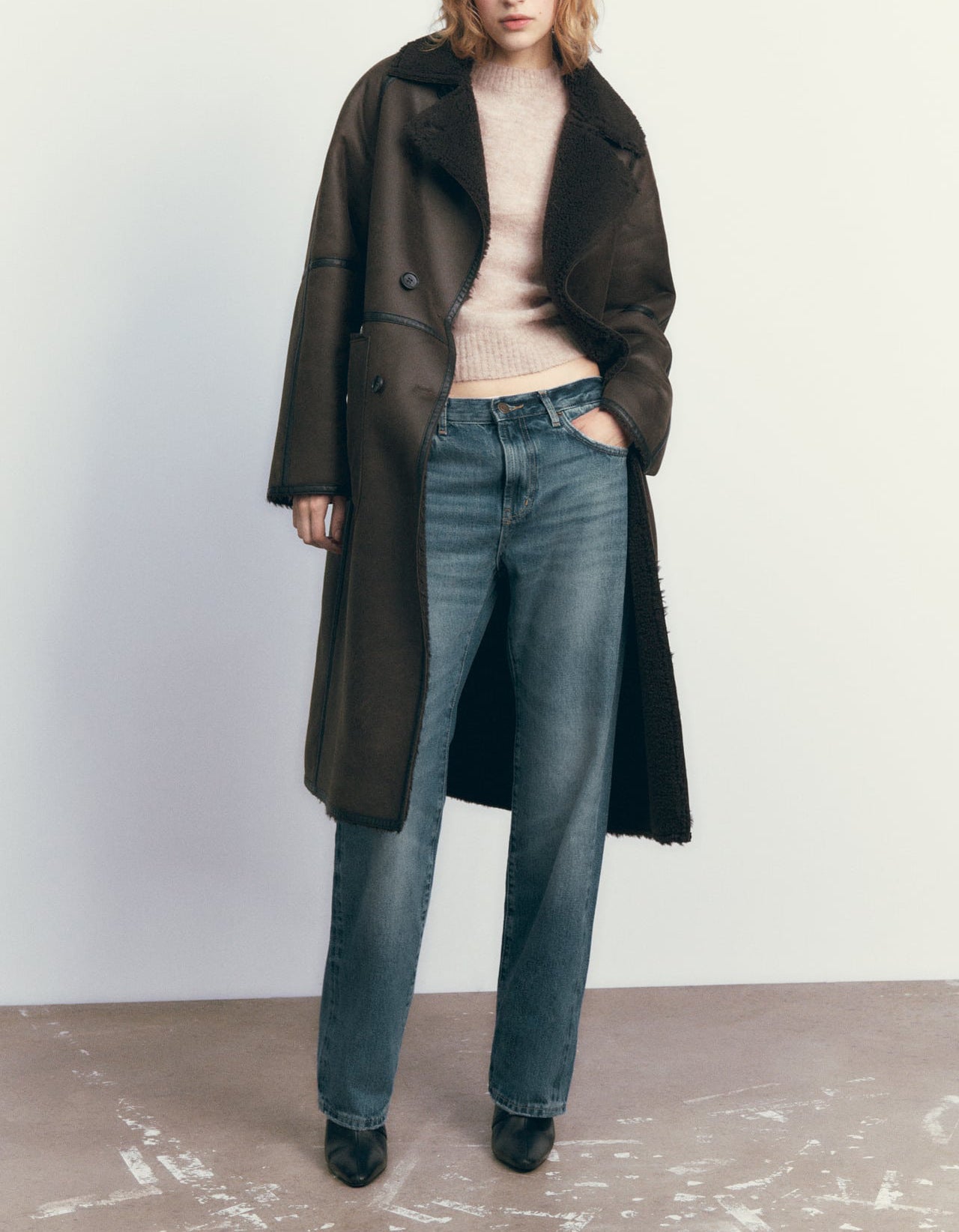 Person wearing a long dark coat and blue jeans against a plain background