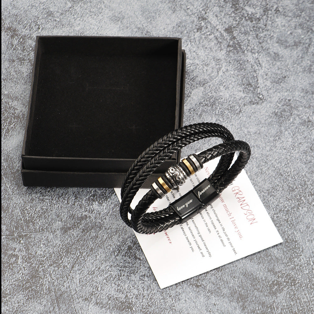 Black braided leather bracelet with metal accents on a gray surface with a black box and paper.