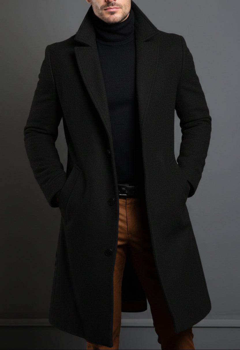 Man wearing a black coat over a black turtleneck and brown pants against a gray background