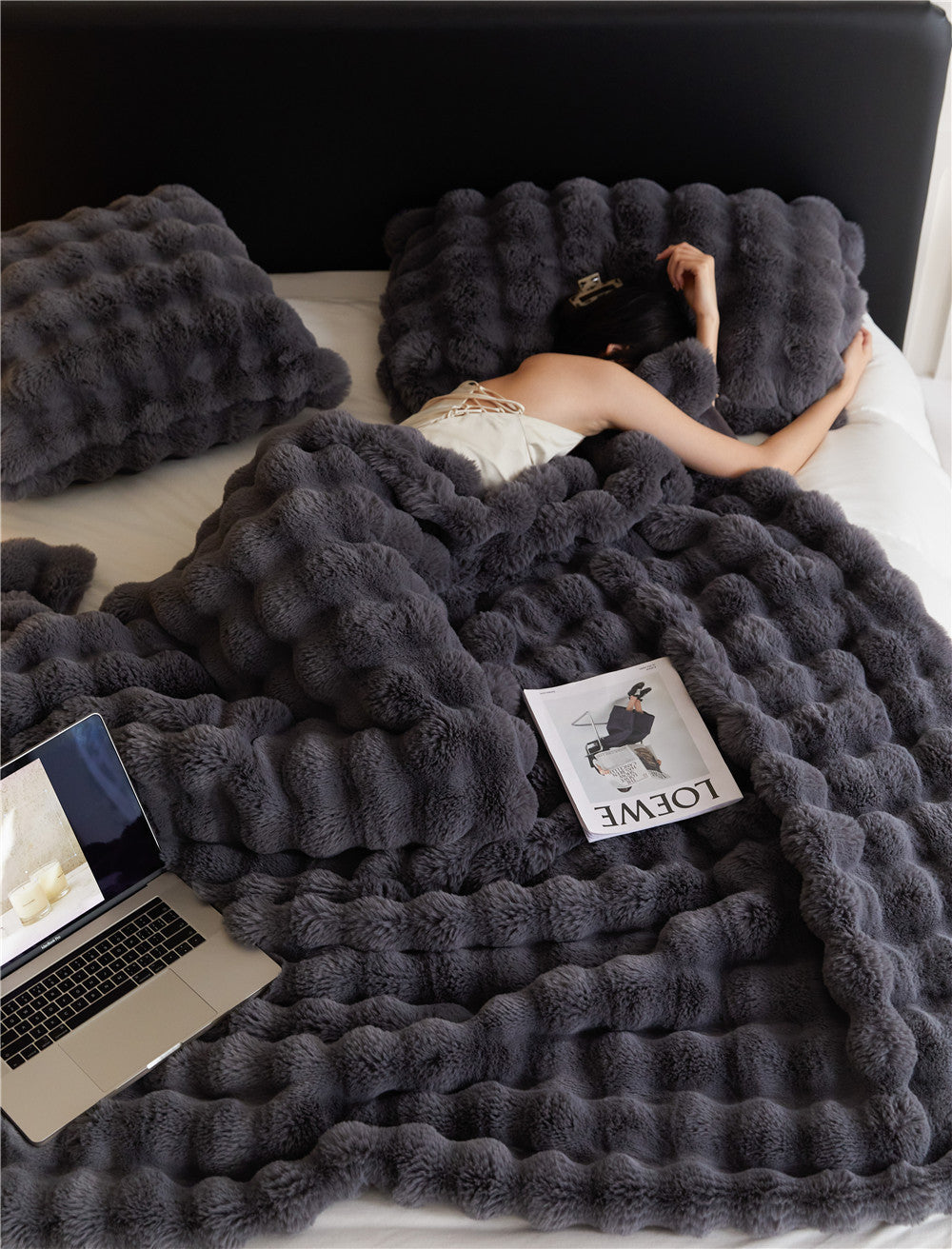 Person lying on a bed under a fluffy gray blanket with a laptop and magazine.