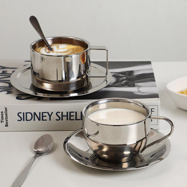 Two silver coffee cups with saucers on a white surface, one filled with a creamy beverage.