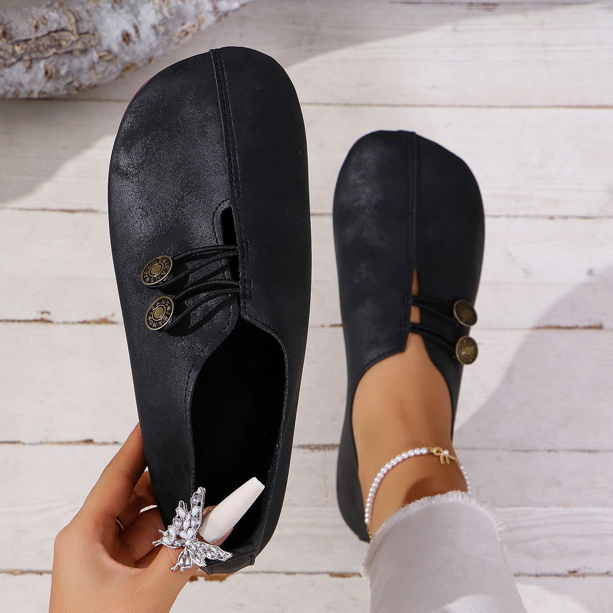 Black flat shoes with decorative buttons worn by a person on a wooden floor.