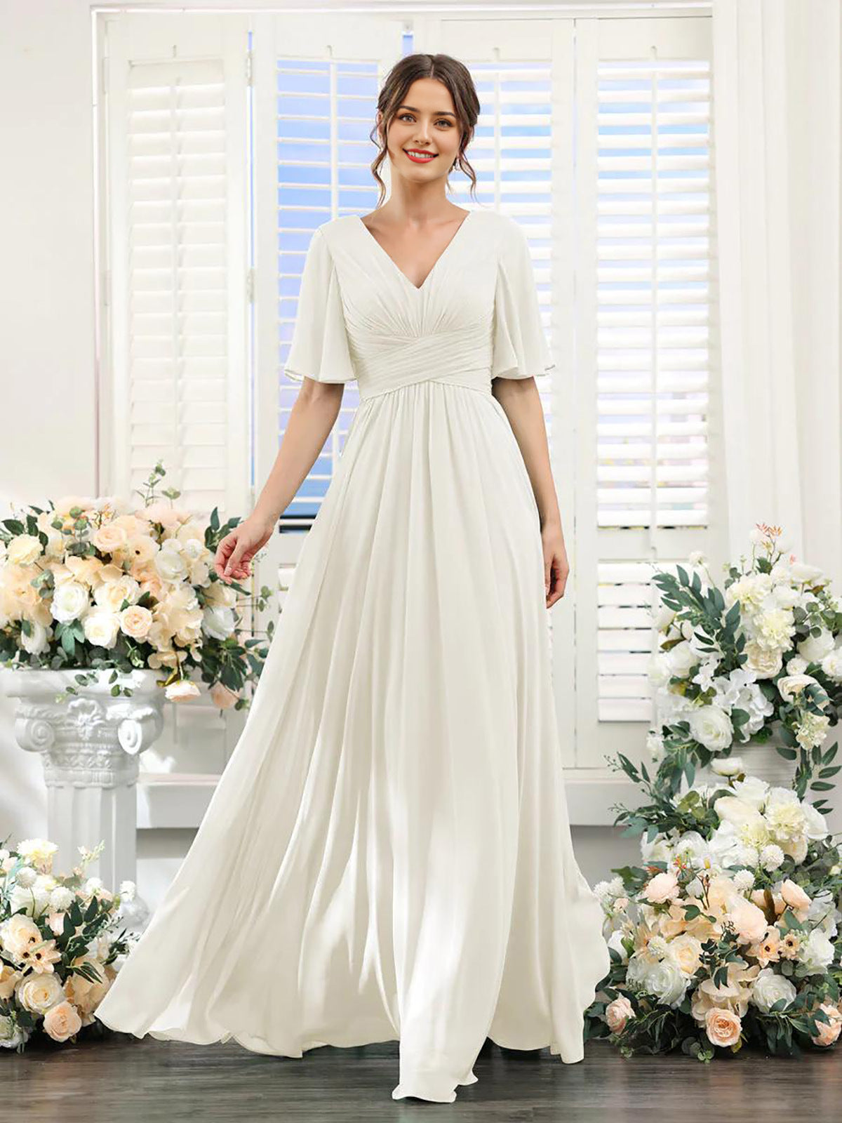 Woman in a white dress standing in front of floral decorations with a window in the background