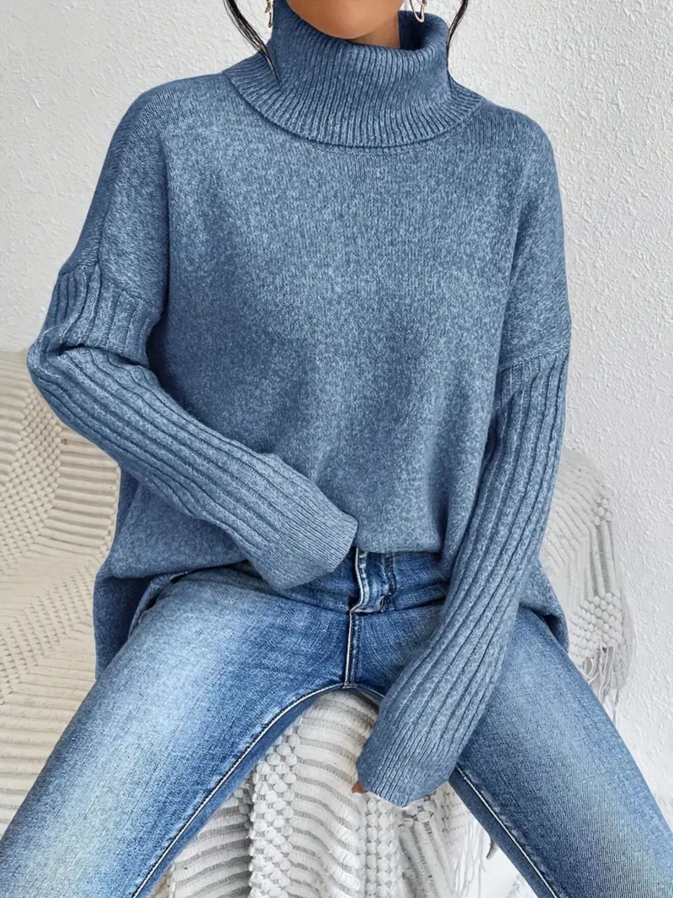 Person wearing a blue turtleneck sweater and jeans sitting on a white chair.