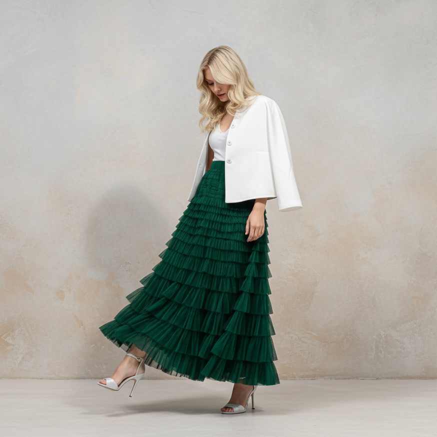 A woman models a long, dark green tiered tulle maxi skirt paired with a white cropped jacket and white heels, standing against a textured beige studio wall.