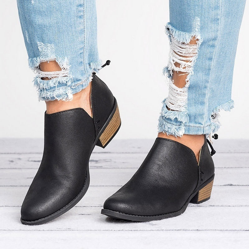 Black ankle boots worn with ripped jeans on a light wooden floor.