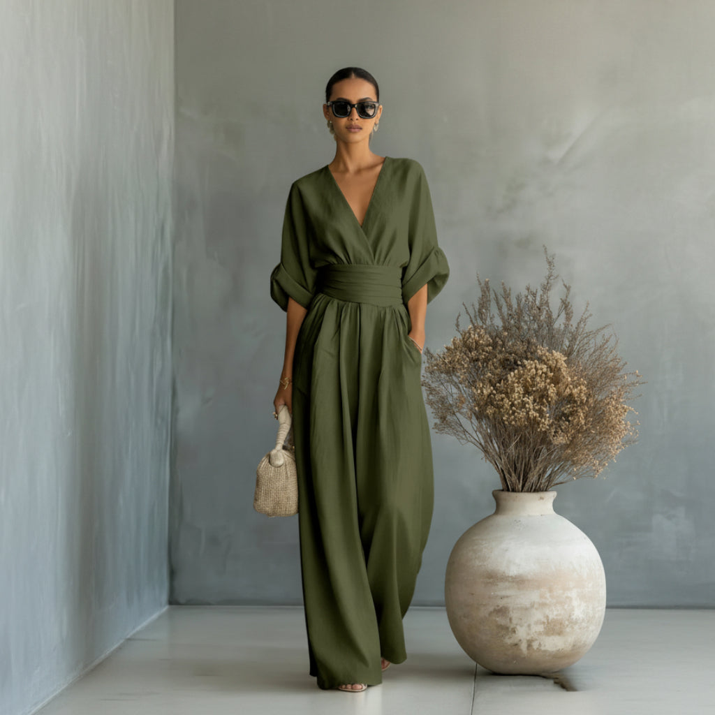Woman wearing a green jumpsuit standing next to a vase with dried flowers against a gray wall.