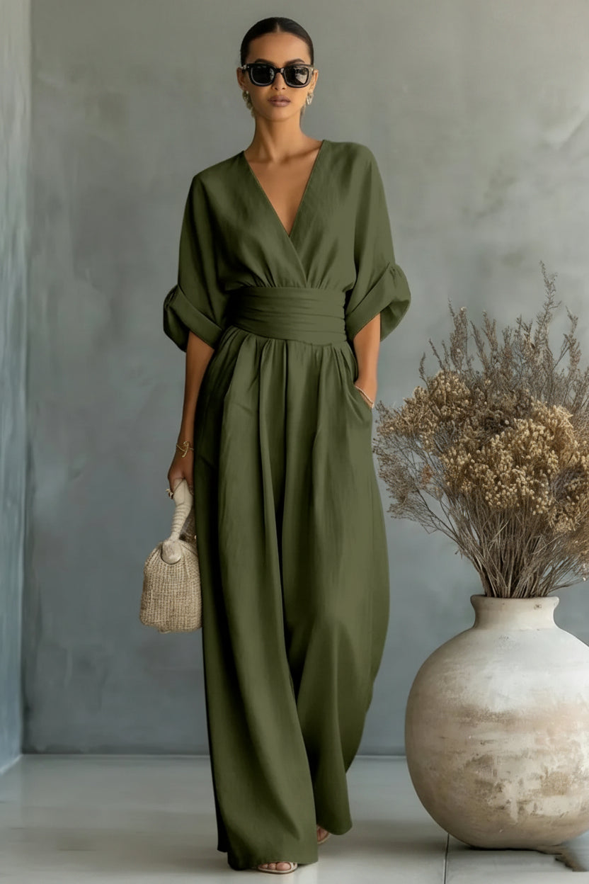 Woman wearing a green jumpsuit standing next to a vase with dried flowers against a gray wall.