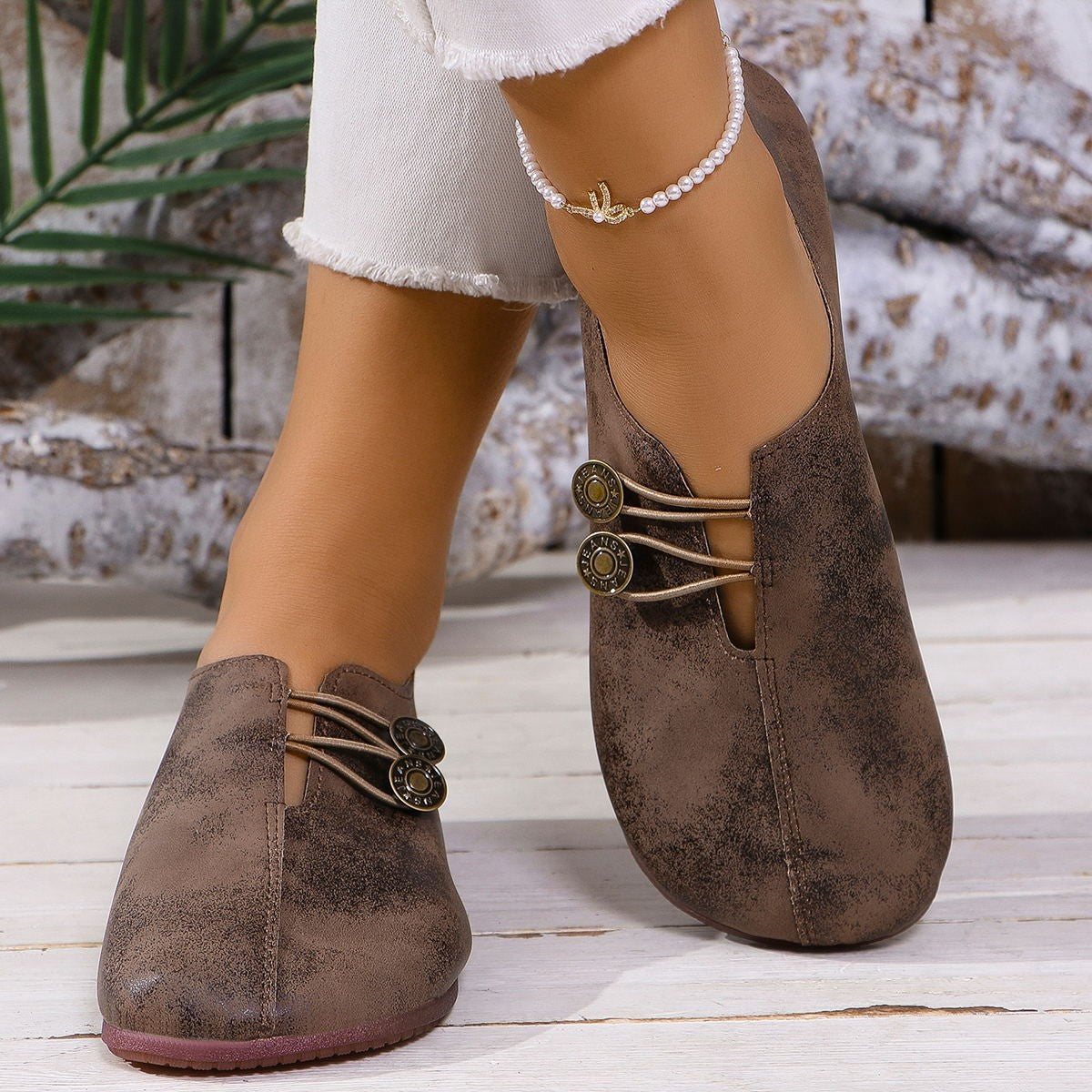 Brown slip-on shoes with decorative straps worn by a person on a wooden floor.