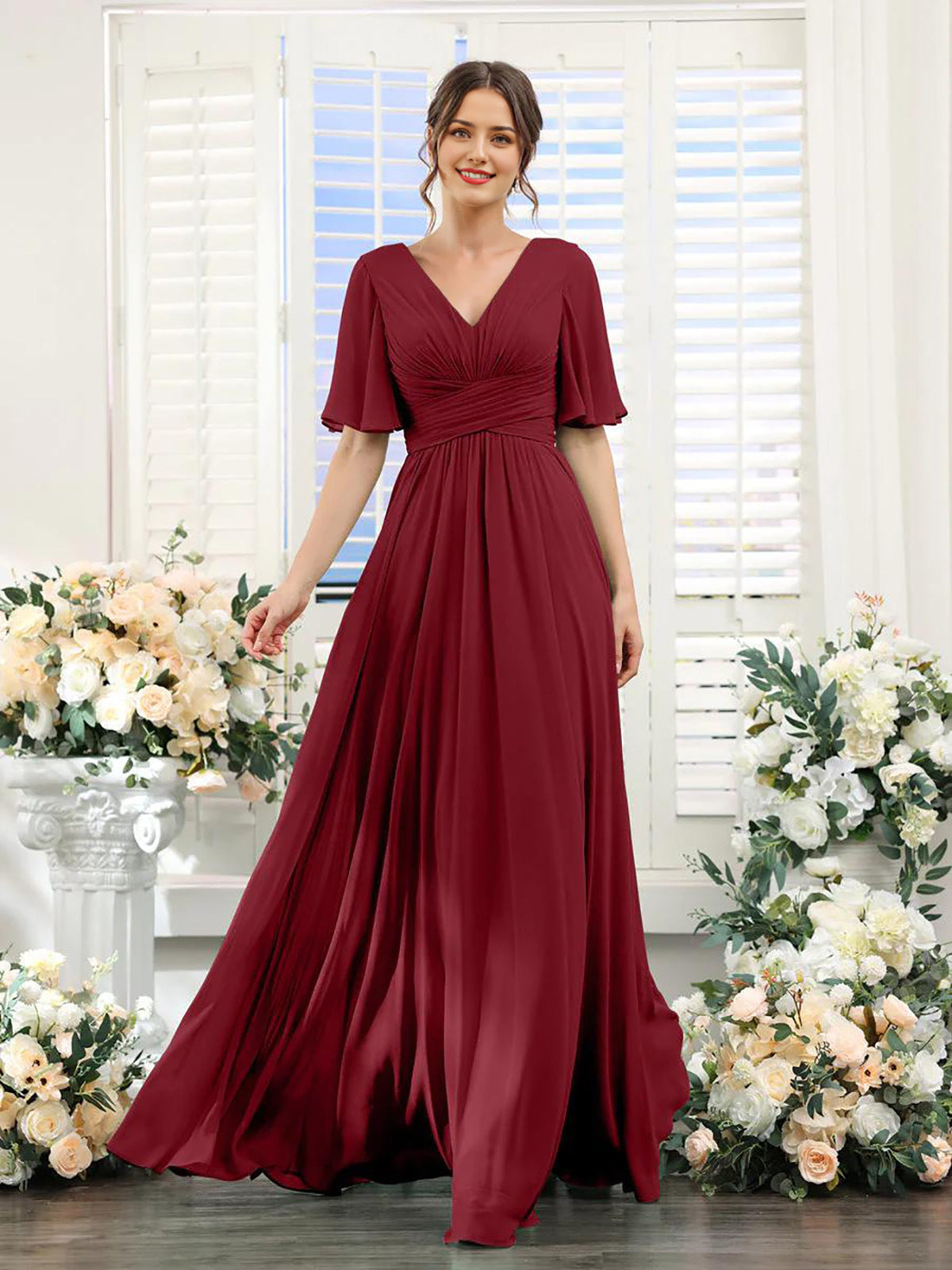 Woman wearing a burgundy dress standing in front of floral decorations and a window with blinds.