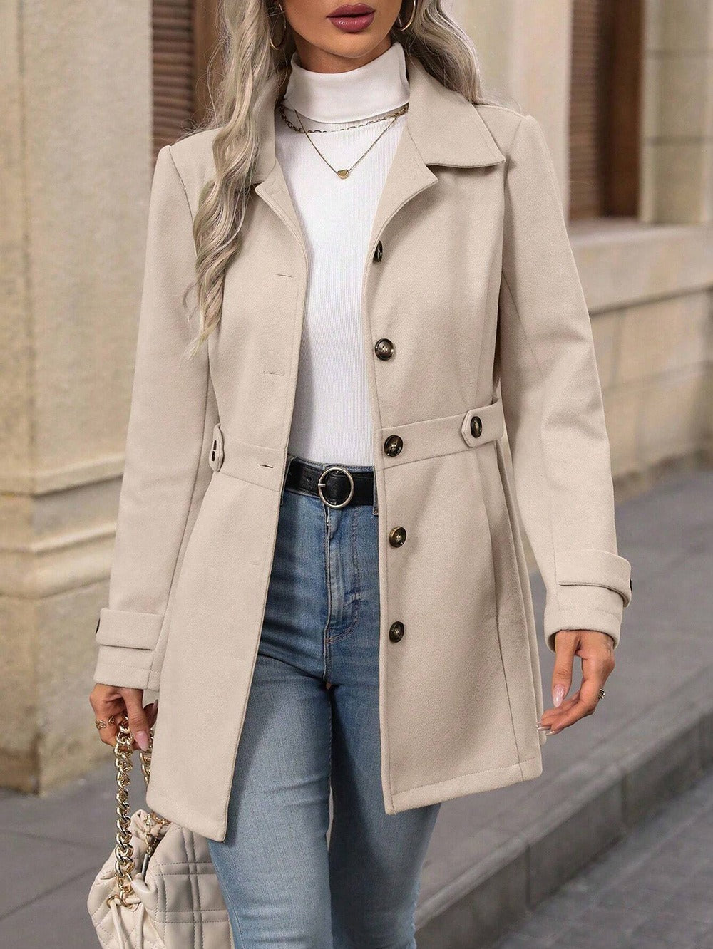 Woman wearing a beige coat, white turtleneck, blue jeans, and carrying a handbag on a city street.