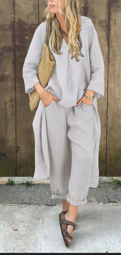 Woman wearing a light gray outfit with wide-leg pants and a long cardigan, standing against a wooden wall.