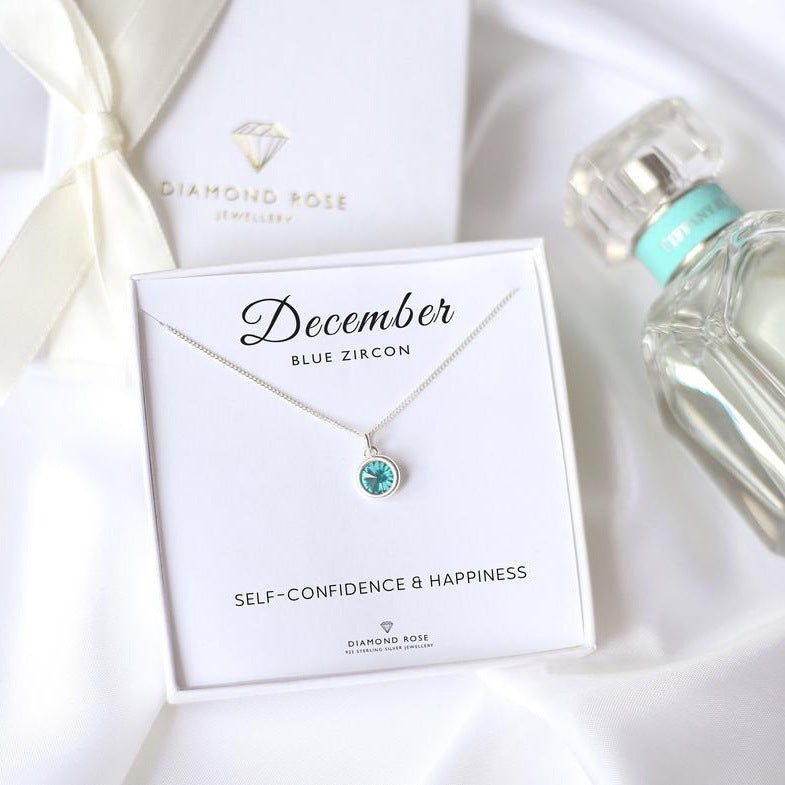 Blue zircon necklace on a card with 'December' and 'Diamond Rose' branding, next to a bottle of perfume.