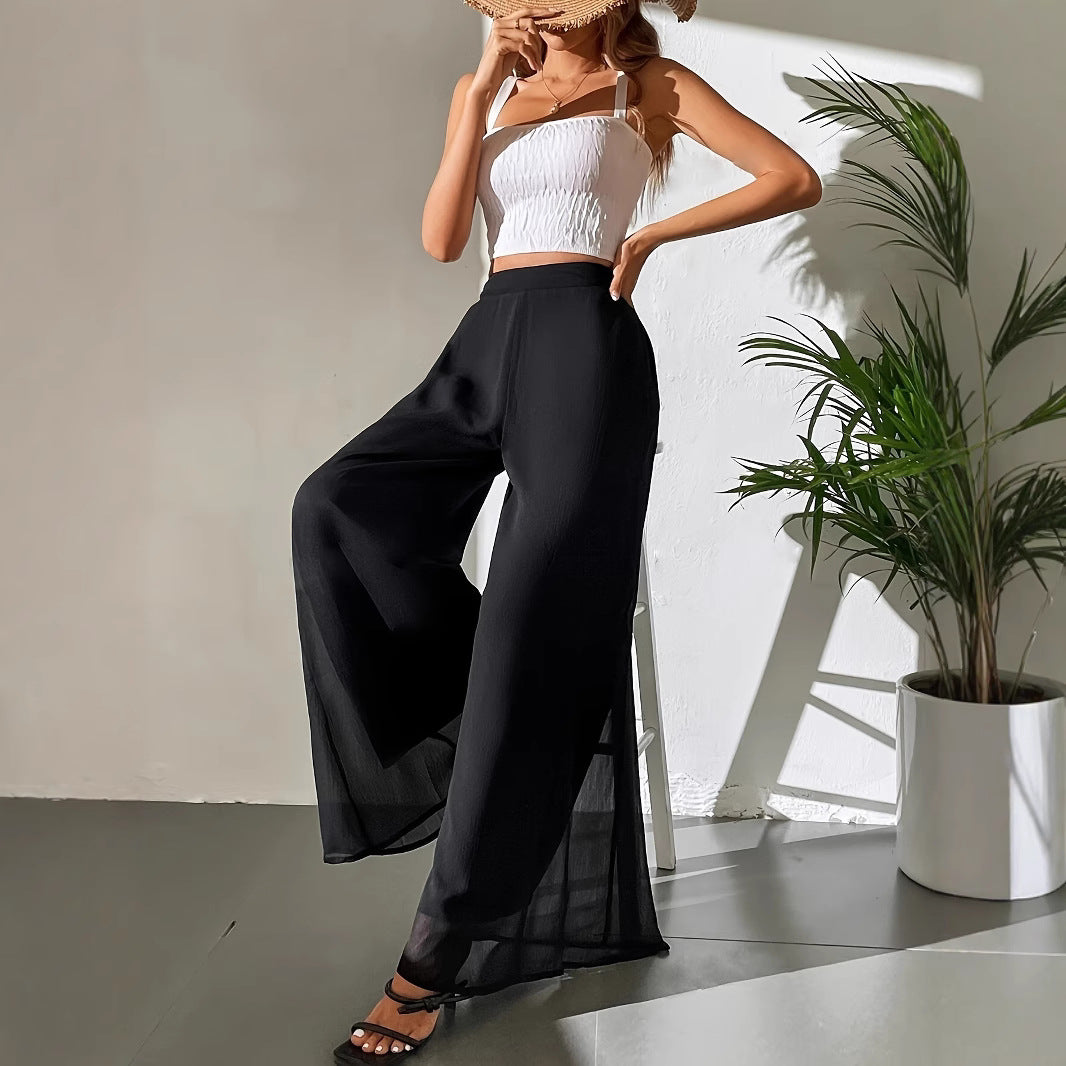 Woman wearing a white top and black wide-leg pants standing next to a plant indoors.