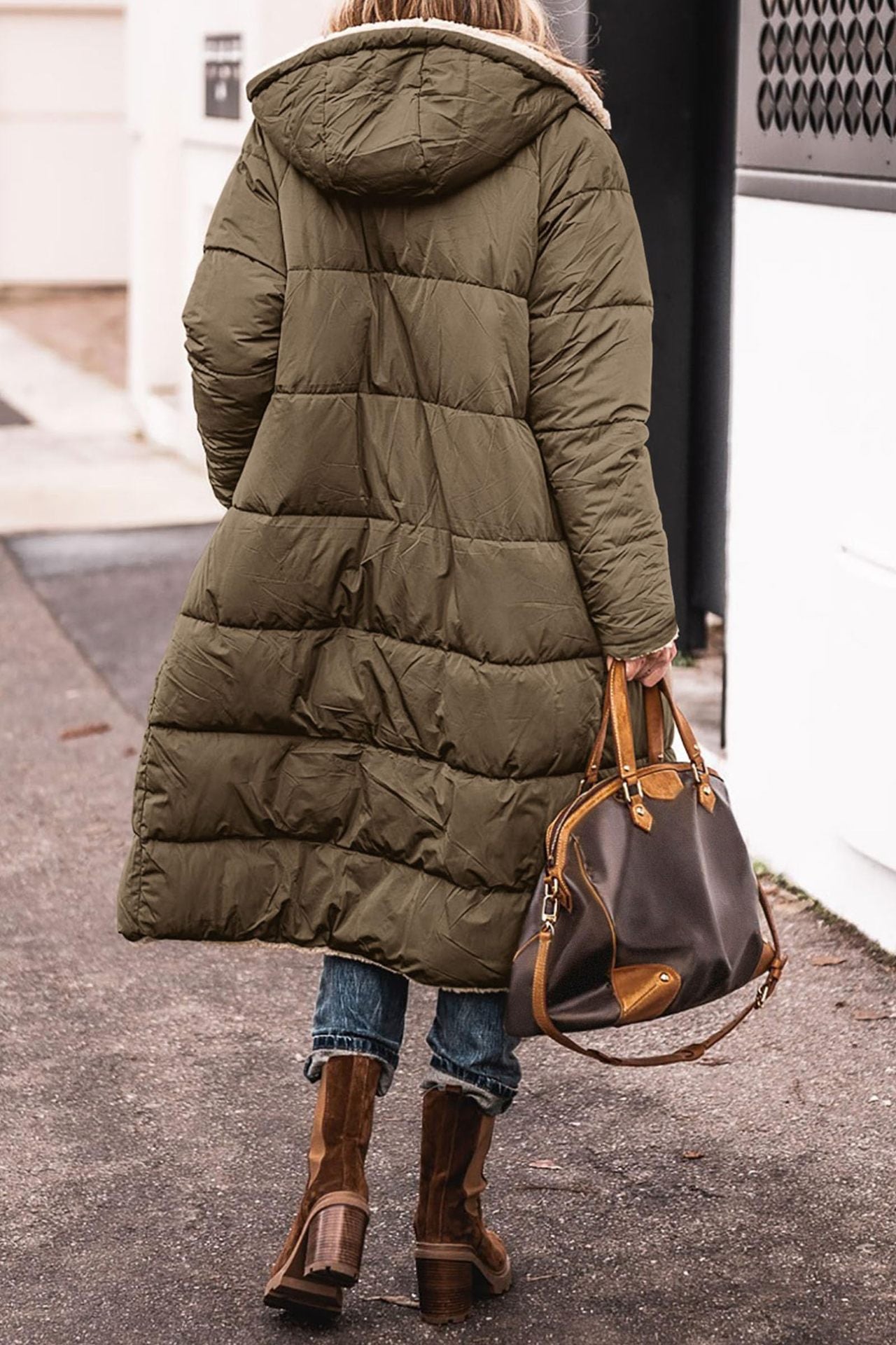 Person wearing a long olive green puffer coat with a hood, holding a brown leather handbag on a sidewalk.