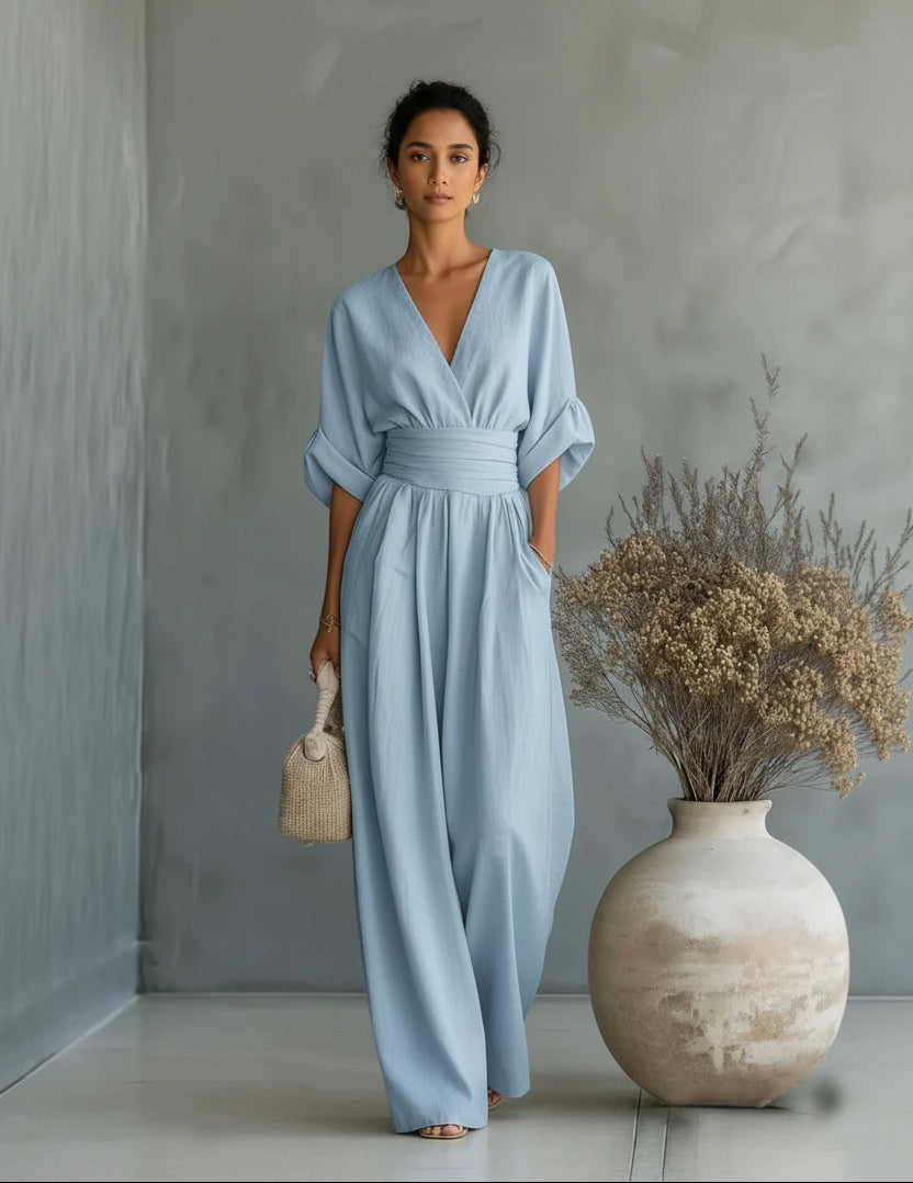 Woman in a light blue dress standing next to a vase with dried flowers against a gray wall.