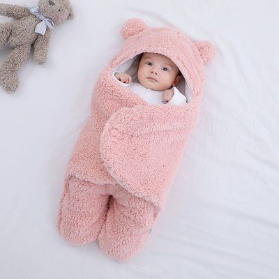 Baby wrapped in a pink fluffy blanket with bear ears, lying on a white surface with a teddy bear beside.