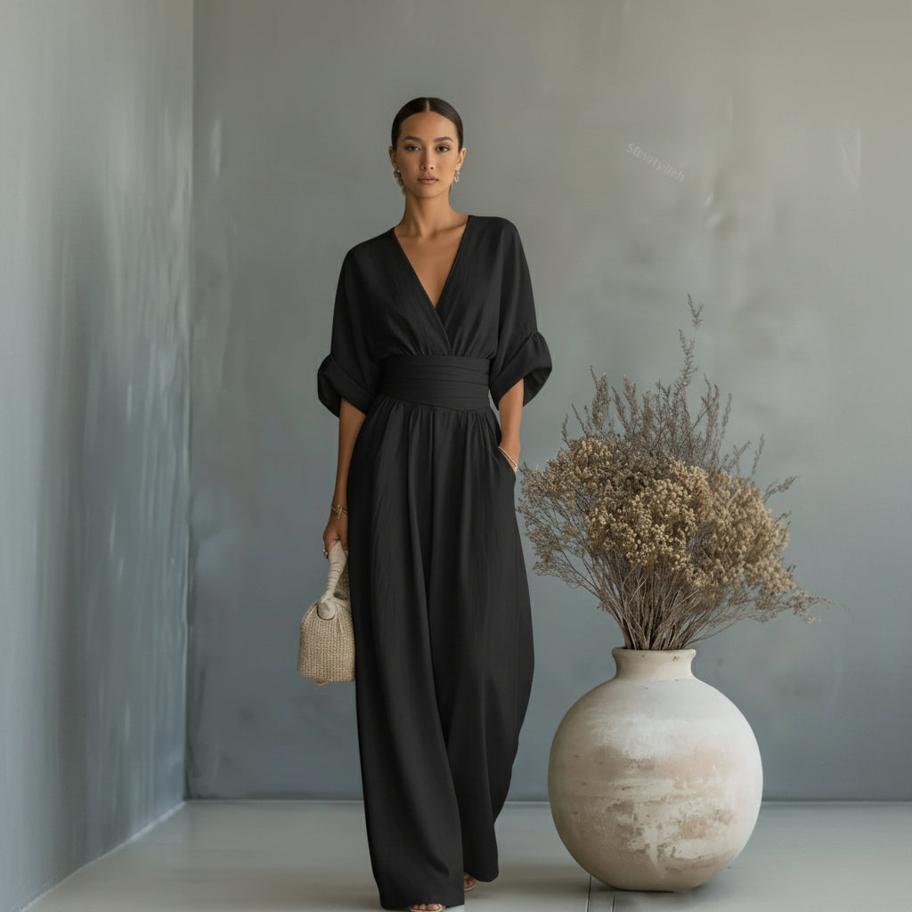 Woman in a black jumpsuit standing next to a textured vase with dried plants against a gray wall.