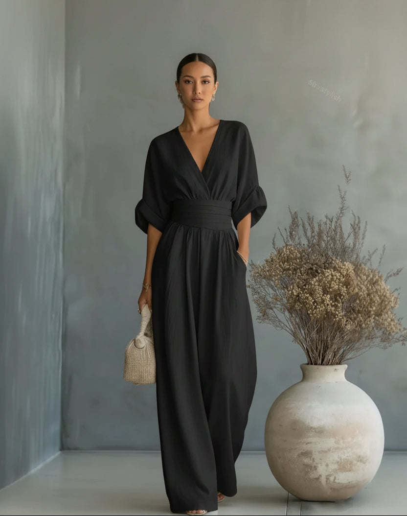 Woman in a black jumpsuit standing next to a textured vase with dried plants against a gray wall.