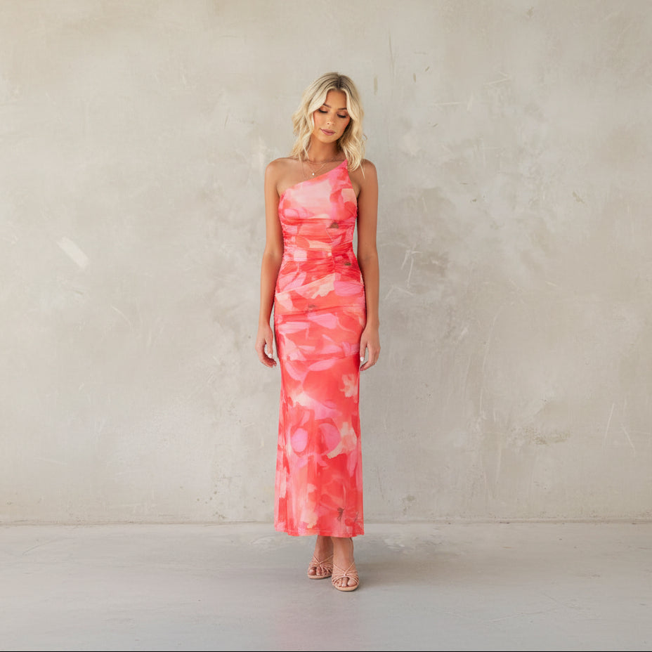 Woman in a pink floral dress standing against a gray concrete wall with a vase of dried plants.