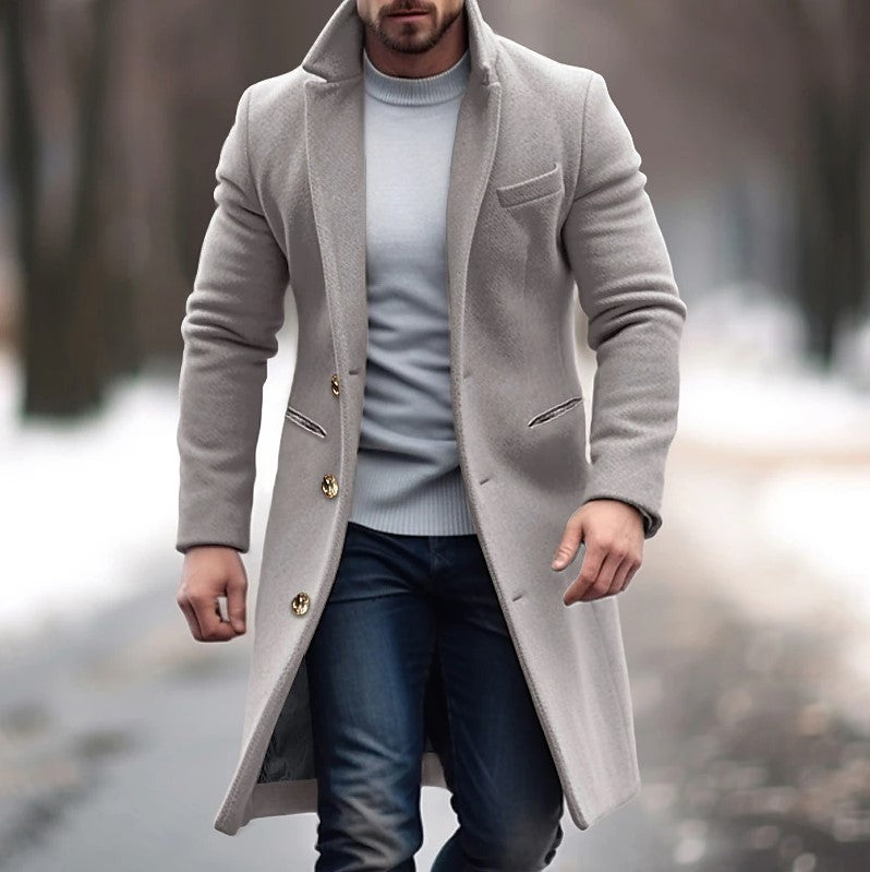 Man wearing a long gray coat over a light blue sweater and dark jeans, standing outdoors.