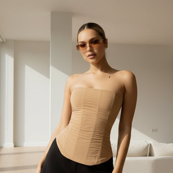 A woman wearing a beige ruched strapless bandeau top and black bottoms, posing on a bed against a plain wall.