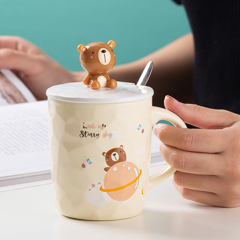 Person holding a mug with a bear design and a small bear figure on top, against a teal background.