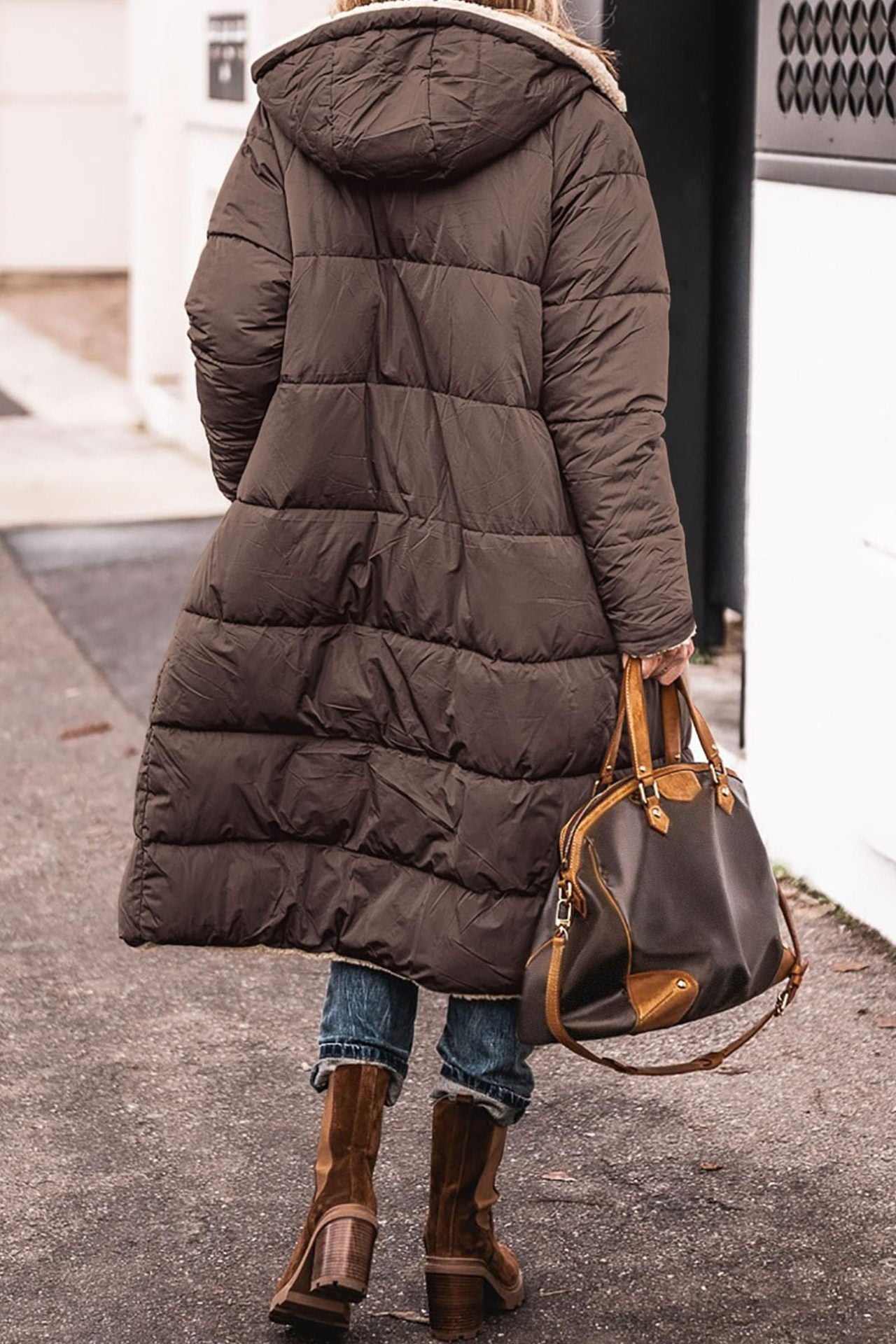 Person wearing a long dark brown puffer coat and holding a matching handbag on a sidewalk.