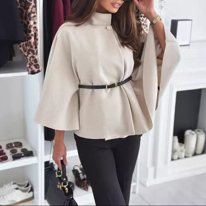 Woman in a beige cape with a black belt in a room with clothing and shoes.