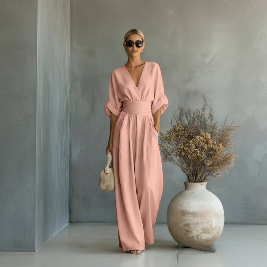 Woman wearing a pink dress standing next to a vase with dried flowers against a gray wall.