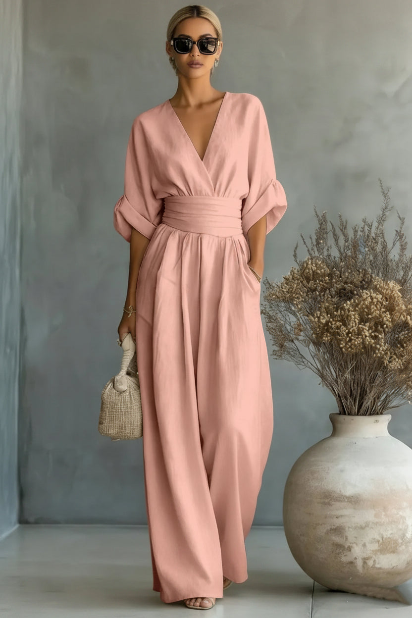 Woman wearing a pink dress standing next to a vase with dried flowers against a gray wall.