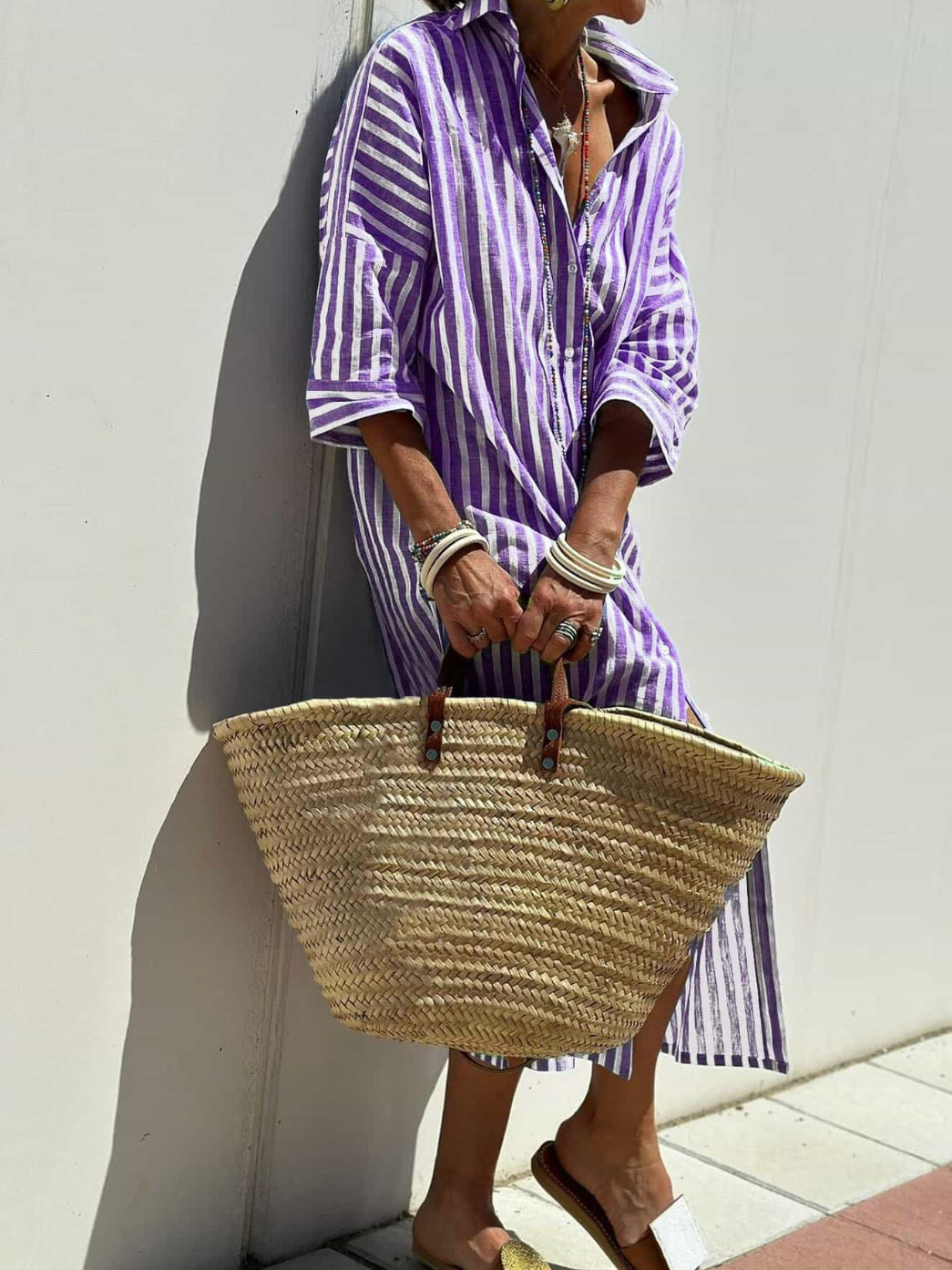 Person wearing a purple and white striped shirt holding a straw bag against a white wall.