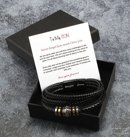 Black braided leather bracelet with gold accents in a black box with a card on a gray textured background