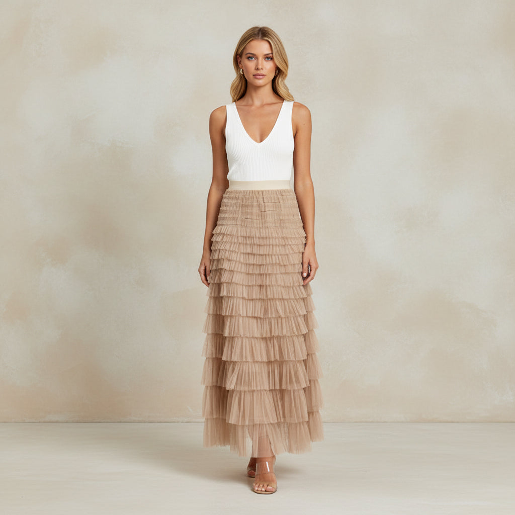 Woman wearing a white top and beige tiered skirt standing against a stone wall.
