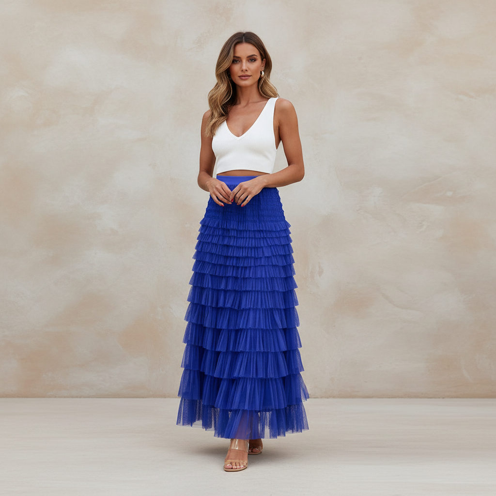 A woman stands in a studio wearing a white sleeveless crop top and a long royal blue tiered tulle maxi skirt, posed against a warm beige textured backdrop.