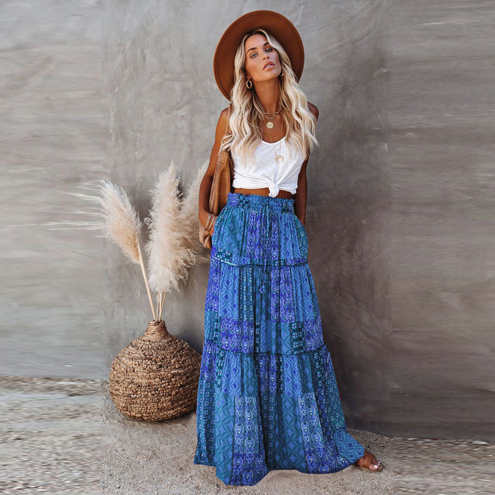 Woman in a blue patterned skirt and brown hat standing next to decorative plants against a gray wall.
