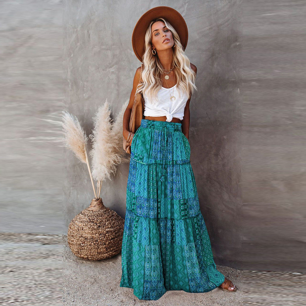 Woman in a white top and green patterned skirt standing next to decorative pampas grass against a neutral wall.