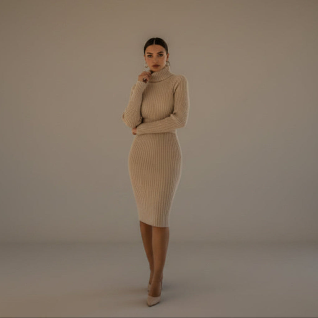 A woman stands in a bright, modern room with large windows, wearing a beige ribbed turtleneck midi dress and nude heels.
