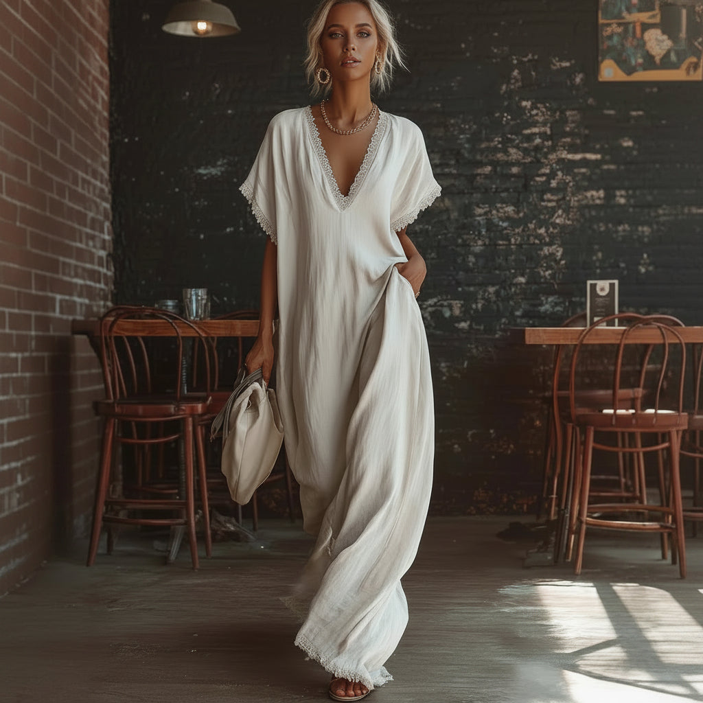 Woman in a flowing white dress standing in a rustic indoor setting with wooden chairs and a brick wall.