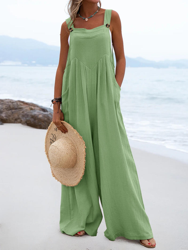 Woman in a green jumpsuit holding a straw hat on a beach.