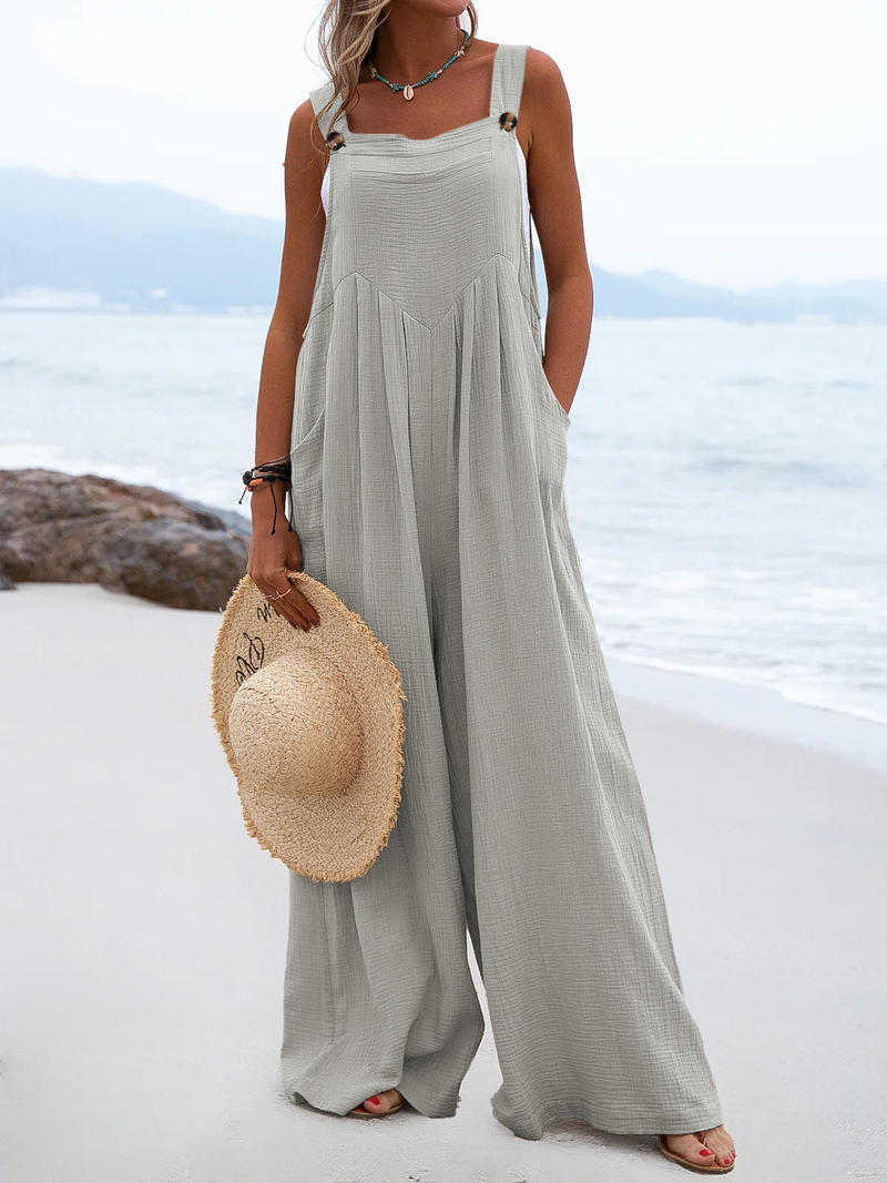 Woman in a light gray jumpsuit holding a straw hat on a beach.